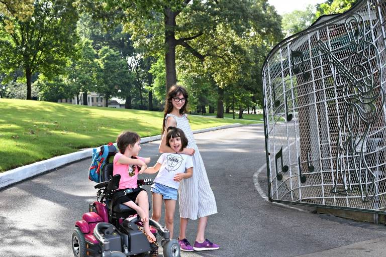 Graceland in Memphis Tennessee. My three girls--all white girls with brown hair--posing infront of the iconic gates in front of Graceland with music notes on the gates. Lamp is in her powerchair and her sisters are standing beside her stricking a funny pose.