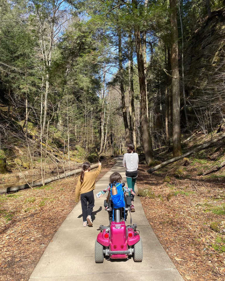 Photo of a path doing through a wooded area. In the middle of the path is the back of a little girl driving a pink wheelchair, with another girl--her sisters--on either side of her, walking along. 