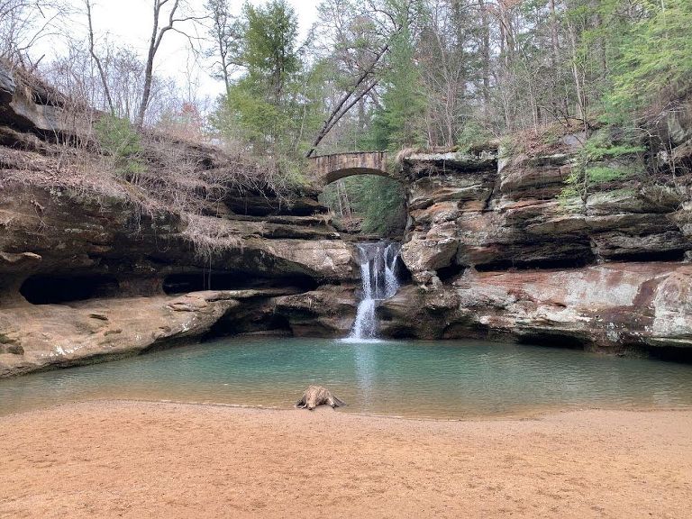 This is a beautiful shot of Old Man's Cave hike in Hocking hills, Ohio. There is a rock wall with a waterfall coming through the middle and a stone bridge going over the waterfall and connecting the two sides of the rock wall. The waterfall goes into a small body of turquoise water and a sandy shore--again all surrounded by rock. 