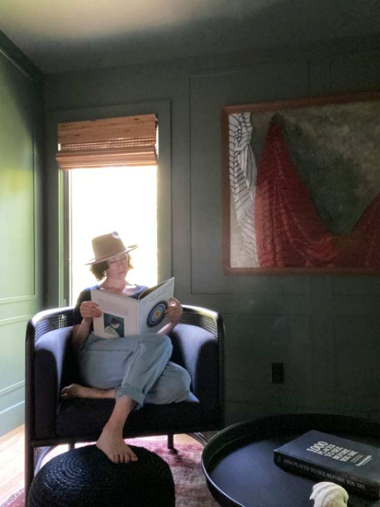 A photo of Amy, a white woman with short dark hair, sitting in a dark green room, on a black chair looking at a book. She's also wearing a hat. 