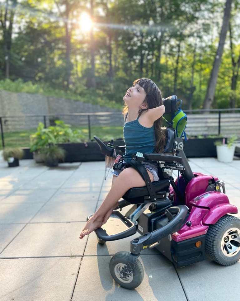 A photo of my daughter Lamp--a white girl with brown hair--sitting in her pink power wheelchair with her arms thrown up in the air, and her head looking up as well, while the sun shines through the trees in the background. 