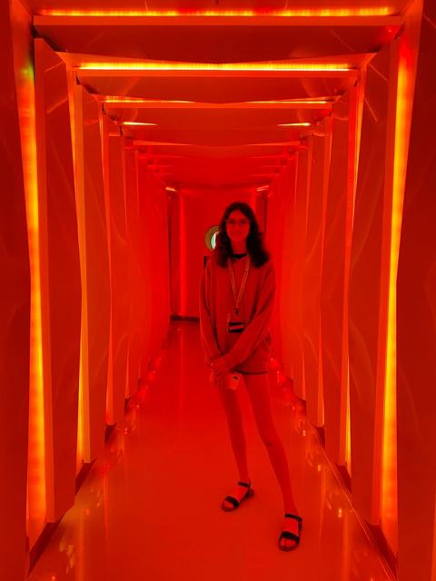 A white girl with dark hair standing in a hallway with red lights lighting the path of a long hallway. Everything is tined red in the photo.