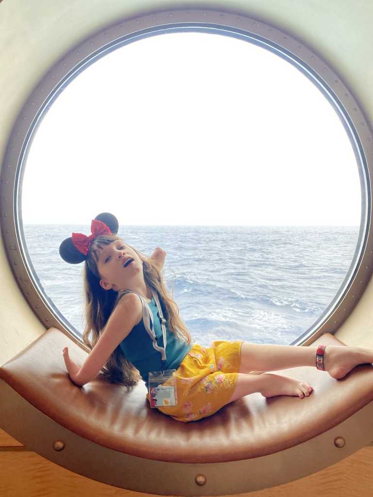 a white girl with long brown hair, who also has limb differences is sitting in front of a round port window. She's leaning back on her arm, in a dramatic gesture of ease with mickey mouse ears on her head.