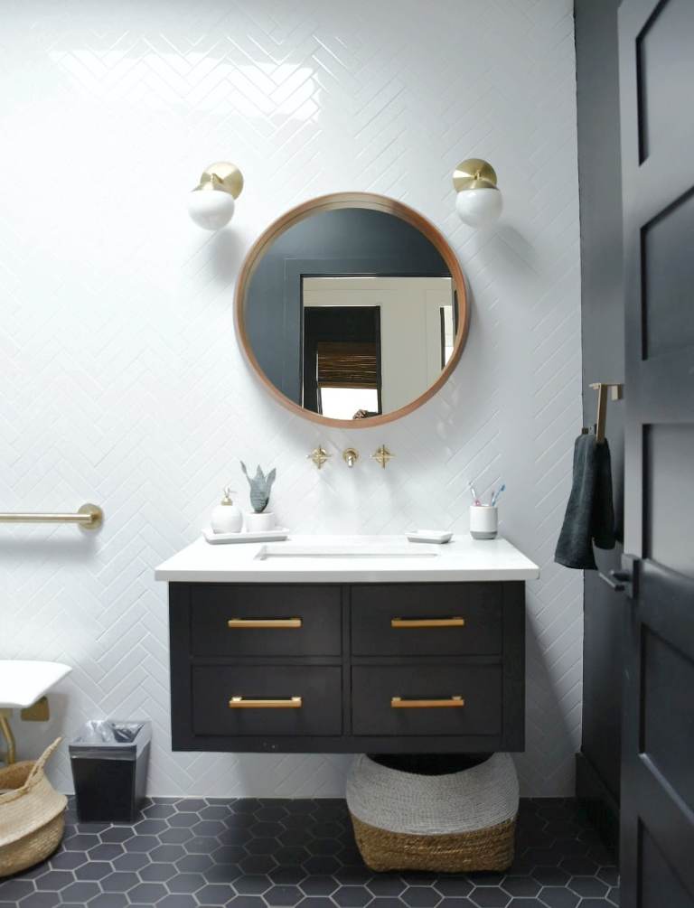 A white tiled wall, with a round wood mirror, and a black wall mounted vanity. There are gold accent lights, as well as gold hard ware and wall mounted faucet. 