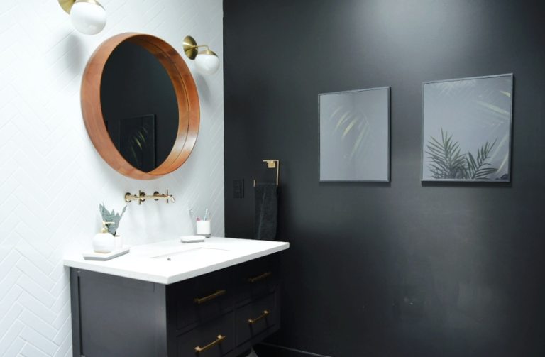 A view of the vanity from the other side, where you see the white tile wall where the mirror and vanity are mounted, next to the black painted wall with two framed photos of palm leaves. 