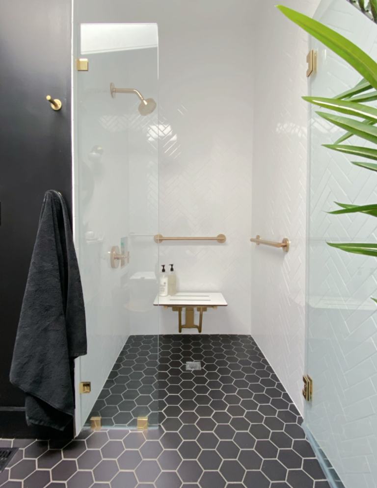 A view inside the black and white bathroom shower, where you see gold grab bars, a fold down bench and a black towel hanging on the hook outside of the shower. 