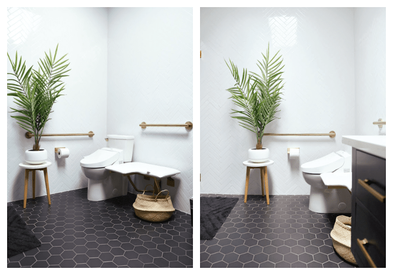 A view of the toilet in the black and white bathroom, with gold grab bars around the toilet, a fold down bench, black hex tile on the floor, and a small side table with a large tropical looking plant on top. 