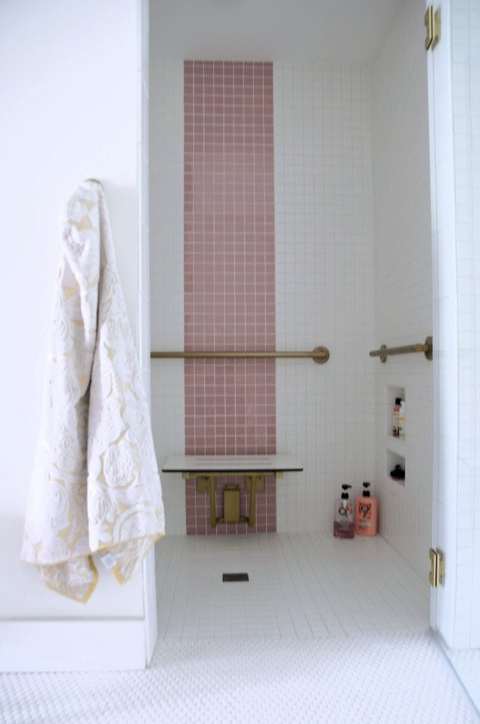 A view of the accessible shower. Again, white tile with a stripe of pink tile going up the center. There are also gold grab bars, a fold down bench and a towel hanging on a hook outside of the shower. 
