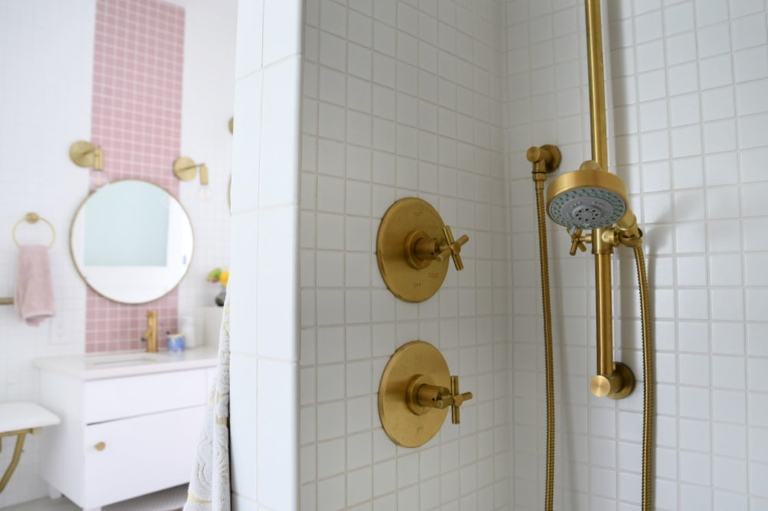 A close up of the gold shower faucets--specifically the gold hand held shower head on a sliding shower bar. You can see the vanity and pink stripes on the wall in the background. 
