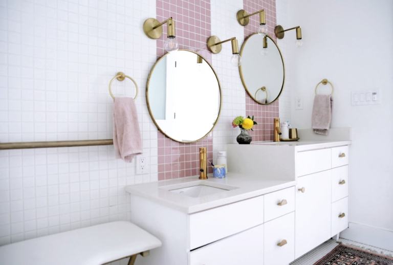 A bathroom vanity with sinks at two different heights, with light pink tile stripes going up the wall behind the round gold mirrors. You can also see gold accent lights, a gold grab bar and towel racks with pink towels on them.
