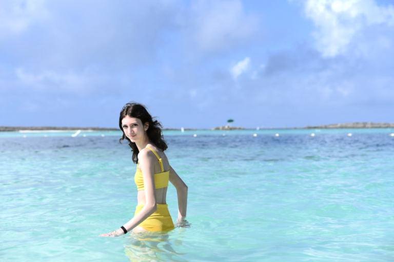 A white girl with dark hair standing waist deep in the ocean in a yellow bikini, looking back at the camera. 
