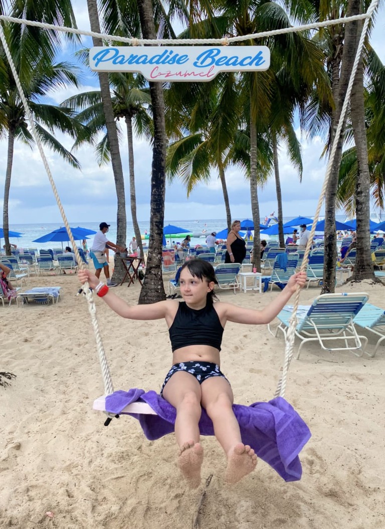 A white girl with dark hair, in a swimsuit on a swing with a sign above her that says, "Paradise Beach, Cozumel." The is a black with palm trees, umbrellas and beach chairs in the background.