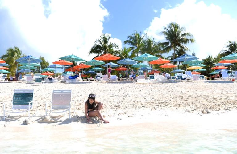 My youngest daughter, a white girl with brown hair, sitting on the beach and digging in the sand with a bunch of beach umbrellas and palm trees in the background.