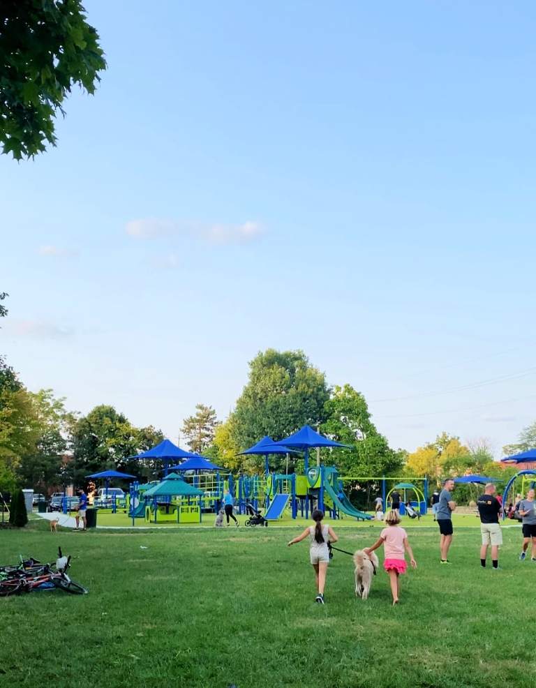 An image of two girls walking a dog as they head toward the finished playground in the background. You can see the blue, green and yellow structure of the playground along with lots of children and adults at the playground in the distance. 